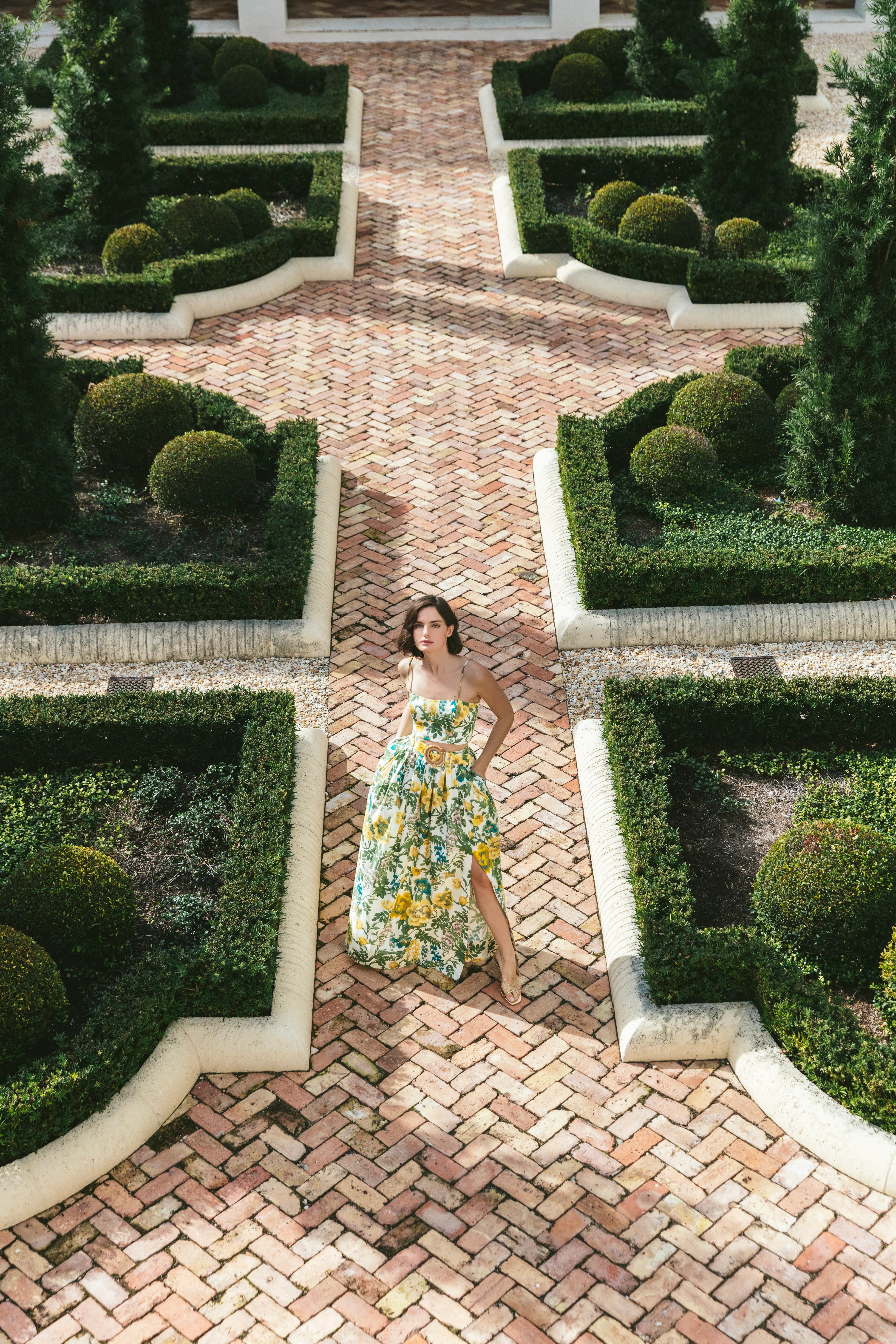 Woman in a floral dress standing in a manicured garden with a brick path.