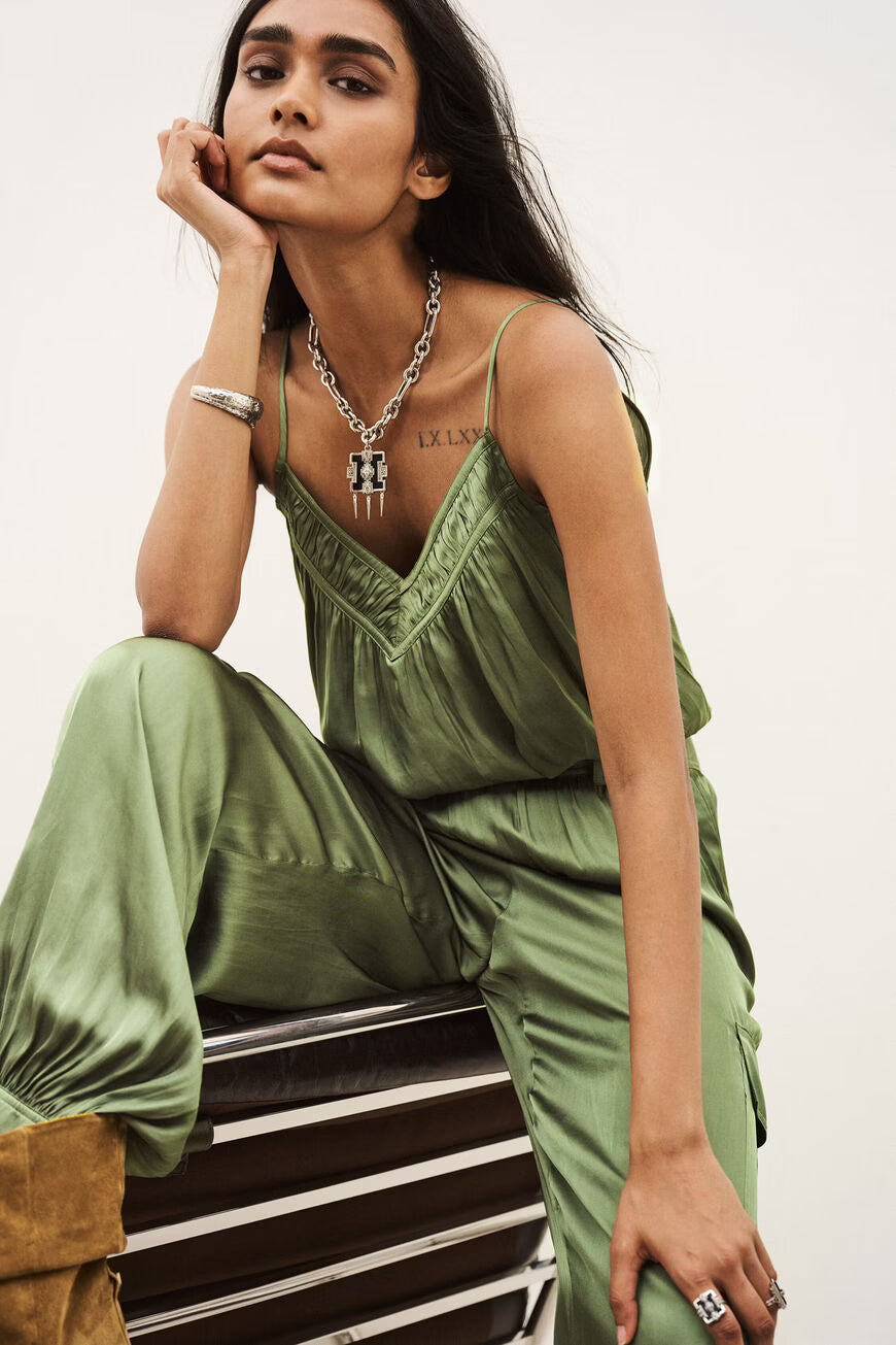 A woman models an olive green satin jumpsuit with matching wide-leg pants, styled with silver jewelry, sitting on a striped wooden bench against a plain white background.