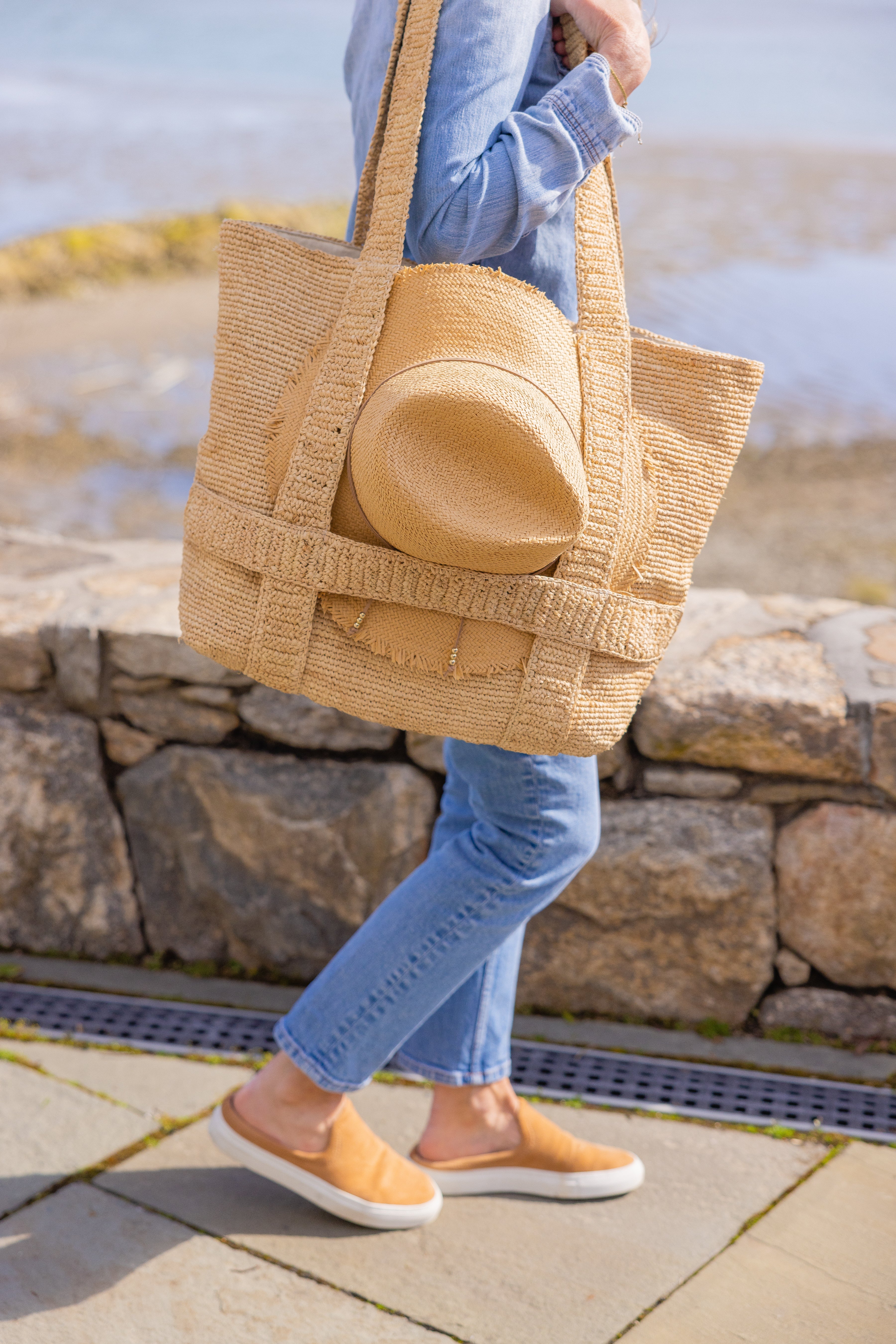 Person holding a large woven bag with a yellow item inside, standing outdoors near a stone wall.