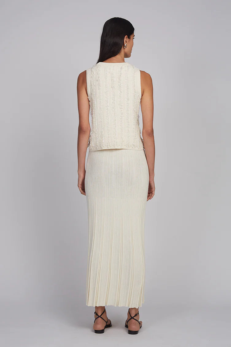 Woman wearing a sleeveless cream top and pleated skirt on a plain background