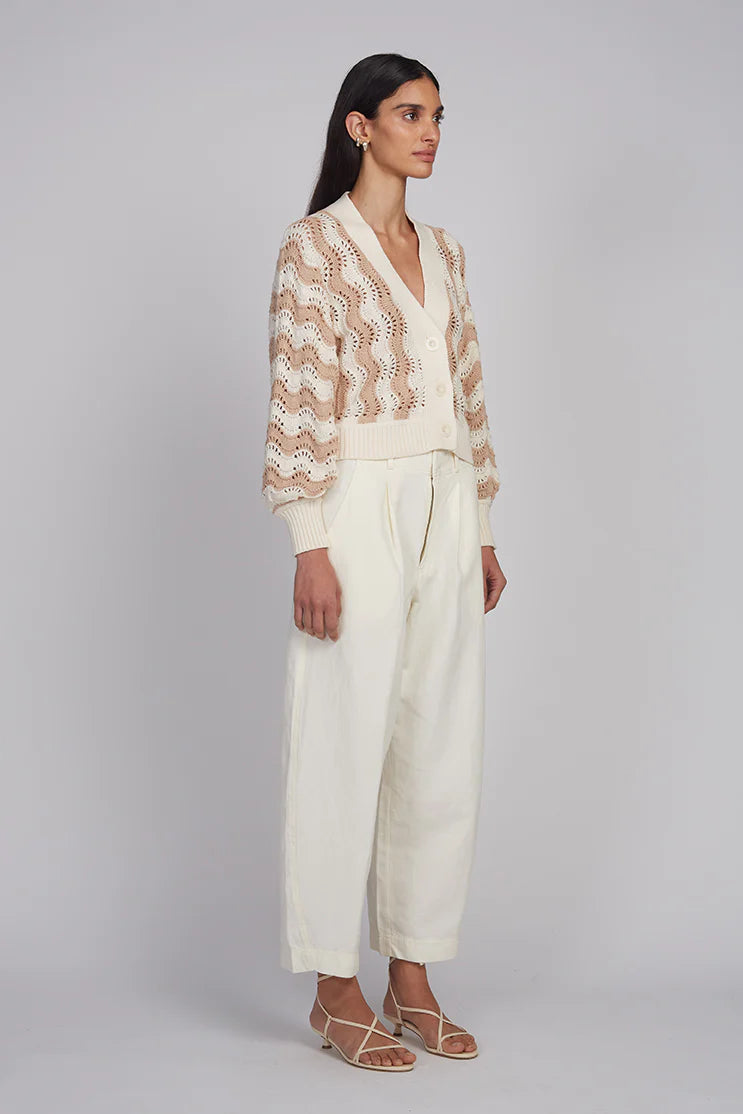 Woman wearing a beige lace cardigan over a white top and pants on a gray background
