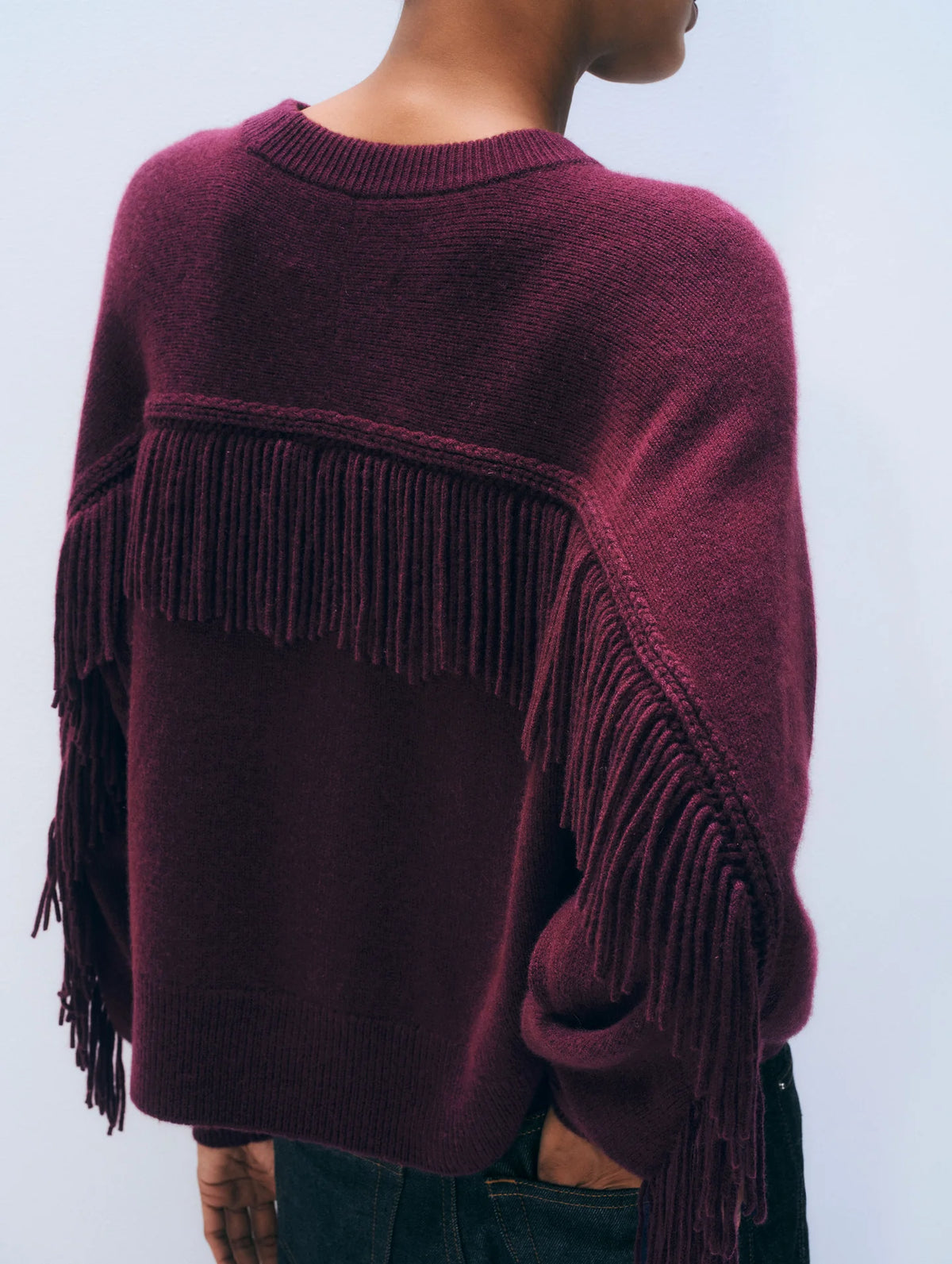 A person wearing a deep burgundy knit sweater with long fringe trim across the back and sleeves, shown from the back against a light gray studio background.