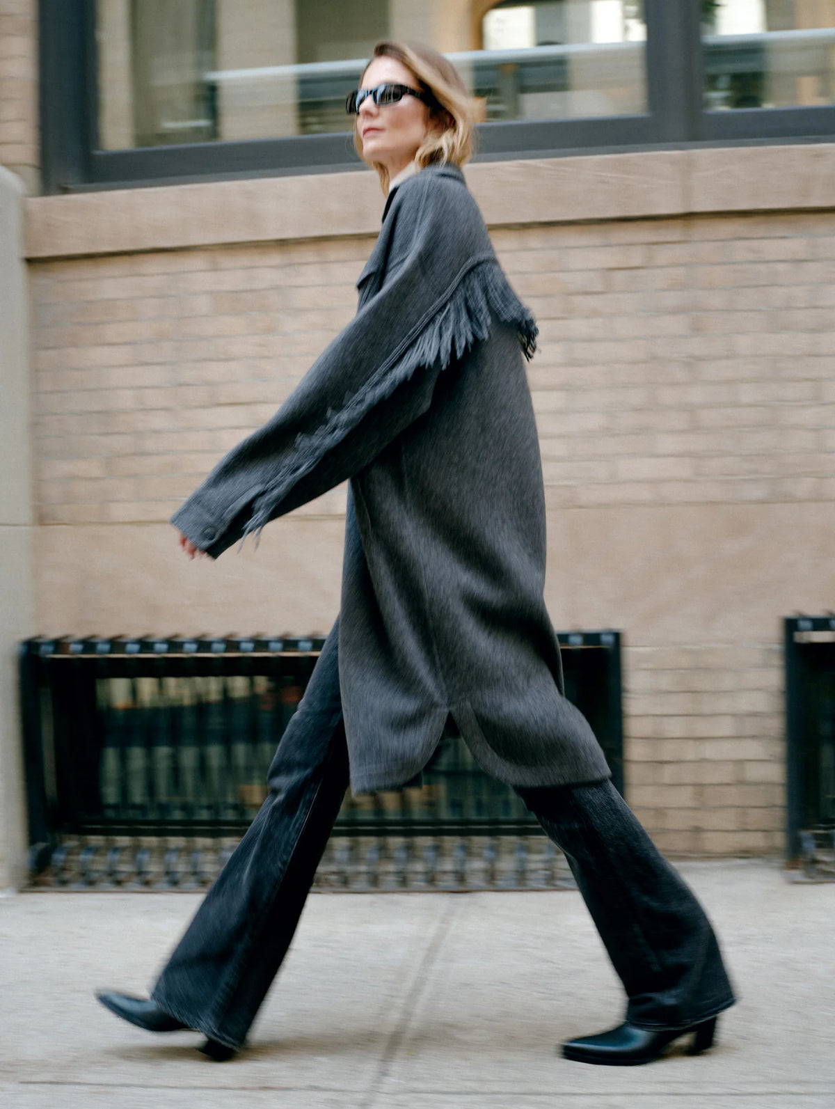 A woman walking outdoors in a city setting, wearing an oversized dark gray knit cardigan with fringe trim, wide-leg dark jeans, black pointed-toe heels, and sunglasses.
