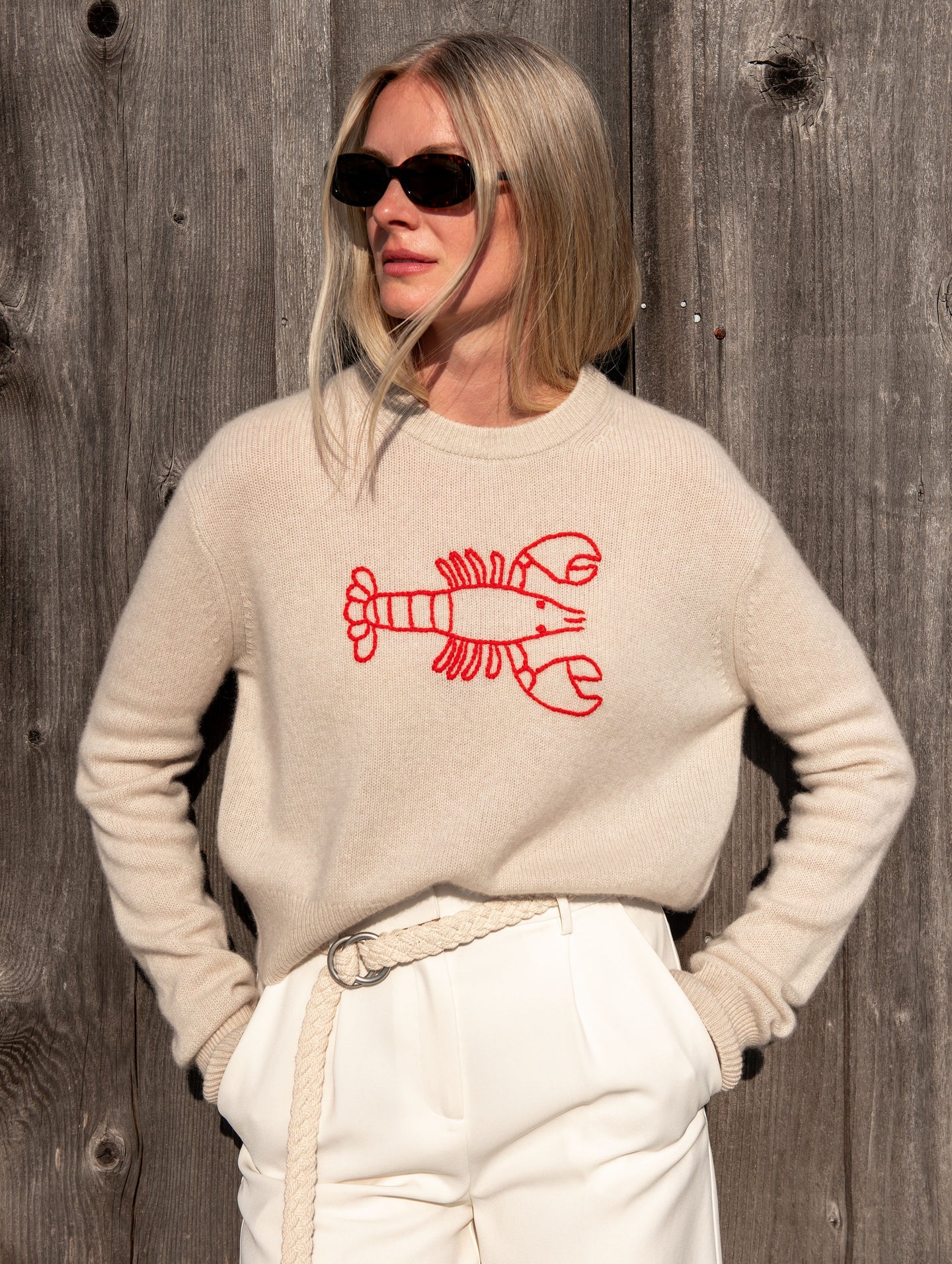 Woman wearing a beige sweater with a red lobster embroidered graphic and cream pants and a cream braided belt on a wooden background