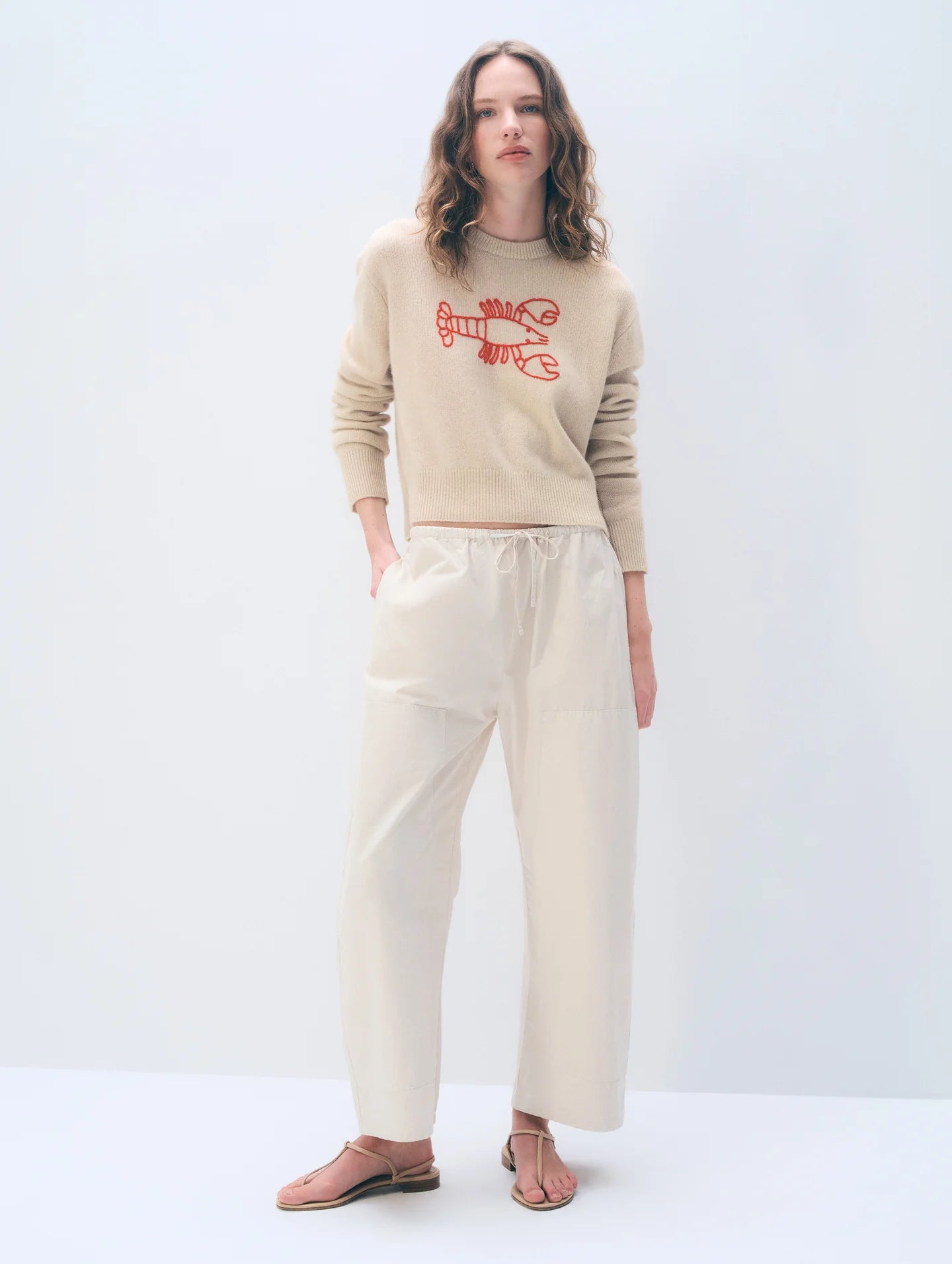 Woman wearing a beige sweater with red text and cream pants on a white background