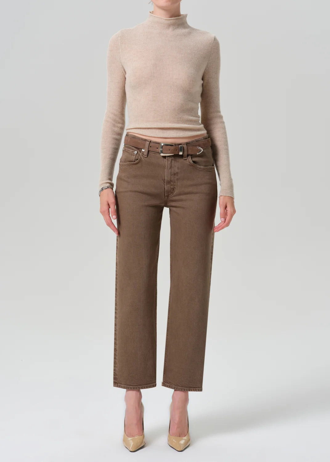 A model wearing a beige ribbed turtleneck sweater and brown wide-leg cropped jeans with a matching belt, paired with beige heels, against a light gray studio background.
