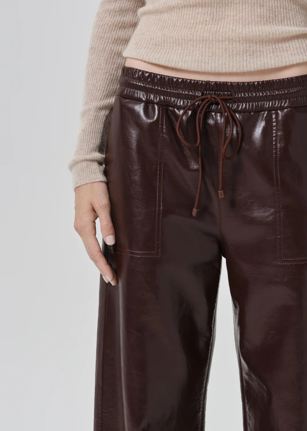 Close-up of a person wearing dark brown faux-leather wide-leg pants with an elastic drawstring waistband, paired with a beige ribbed sweater, against a light gray studio background.