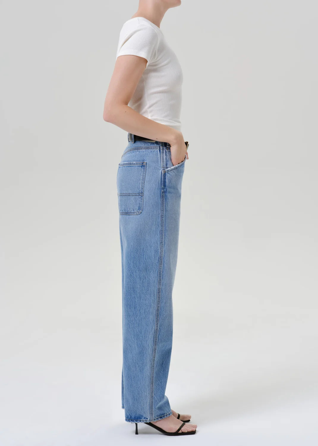 A side-profile view of a model wearing light-wash blue wide-leg jeans, a white short-sleeve t-shirt, and black heeled sandals against a light gray studio background.