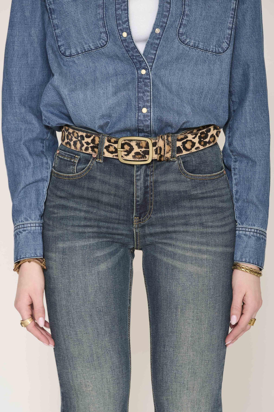 A close-up of a person wearing a leopard-print belt with a gold-tone rectangular buckle, styled with a blue denim shirt and blue jeans against a light beige background.
