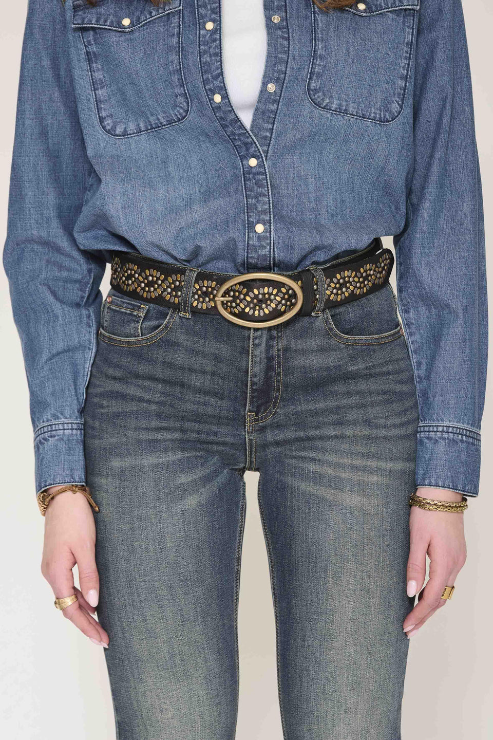A close-up of a person wearing a black studded belt with a large oval gold-tone buckle, paired with blue jeans and a blue denim shirt.