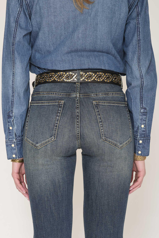 Rear view of a person wearing blue denim jeans with a decorative black belt featuring gold-toned studs and a blue denim shirt tucked in, against a plain light background.