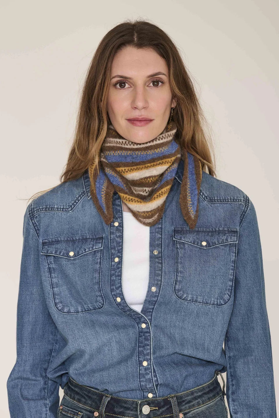 A woman wearing a multi-colored striped knit scarf in shades of blue, brown, and beige, styled with a blue denim shirt over a white top.