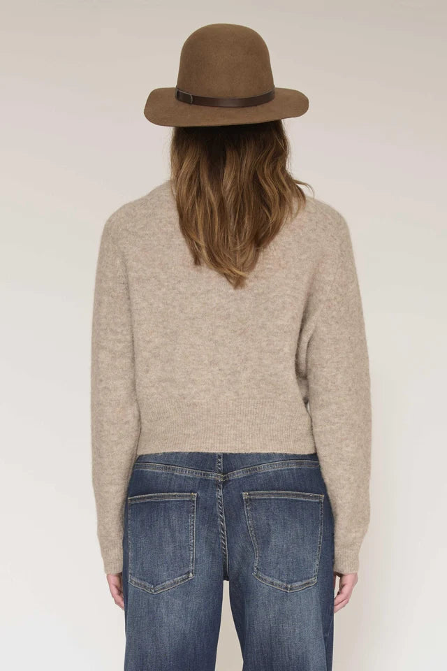 A woman shown from the back wearing a beige knit sweater, blue jeans, and a brown felt hat against a plain light background.