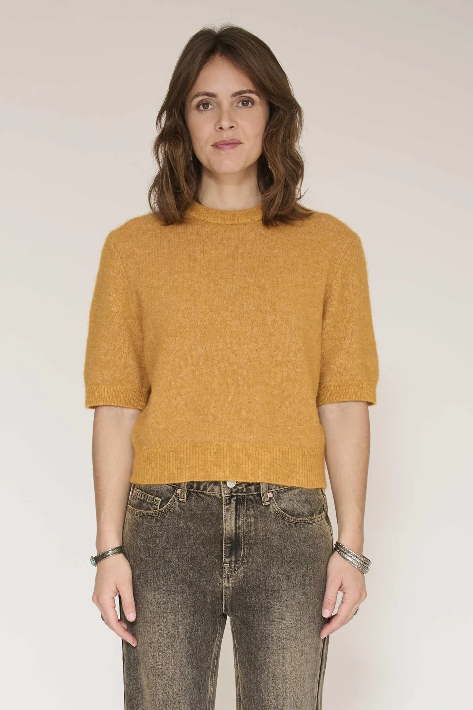 A woman wearing a mustard yellow short-sleeve knit sweater and dark gray jeans, standing against a plain light beige studio background.