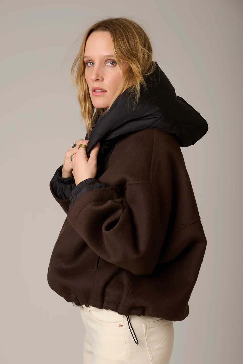 A woman wearing a dark brown oversized hooded jacket with a black puffer-lined hood, paired with off-white pants, photographed against a neutral gray studio background.
