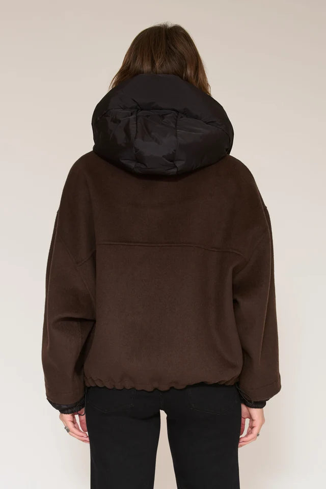 A person viewed from the back wearing a dark brown hooded jacket with a black hood and black pants, standing against a plain light beige studio background.