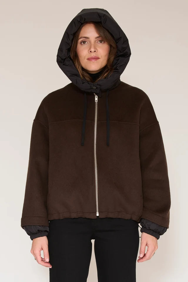 A woman wearing a dark brown zip-up hooded jacket with a black inner hood lining and black cuffs, paired with black pants, against a plain light beige studio background.