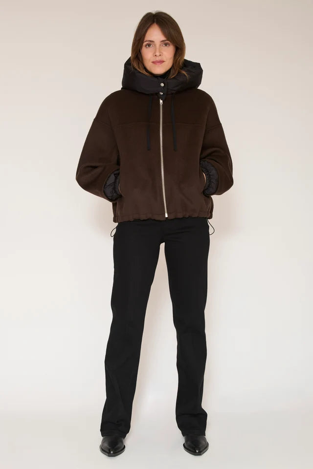 A model stands against a plain light background wearing a dark brown hooded jacket with a black front zipper and black gloves, paired with black pants and black shoes.