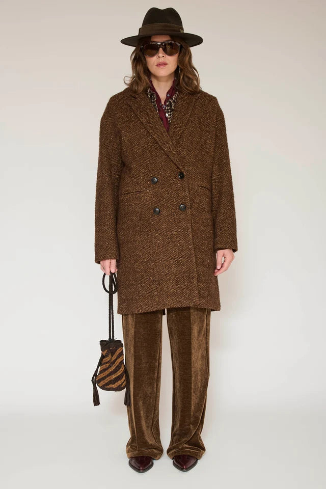 A model wearing a brown double-breasted tweed coat, matching brown velvet trousers, a brown fedora hat, sunglasses, and holding a brown and black striped bucket bag against a plain light background.