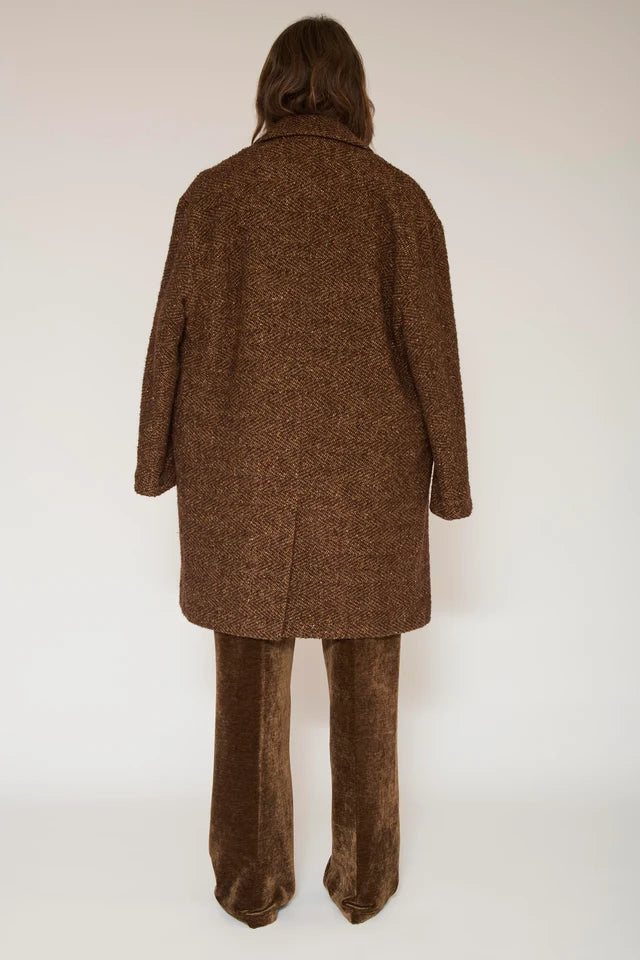 A model viewed from the back wearing a brown textured tweed coat and matching brown wide-leg trousers against a plain off-white studio background.
