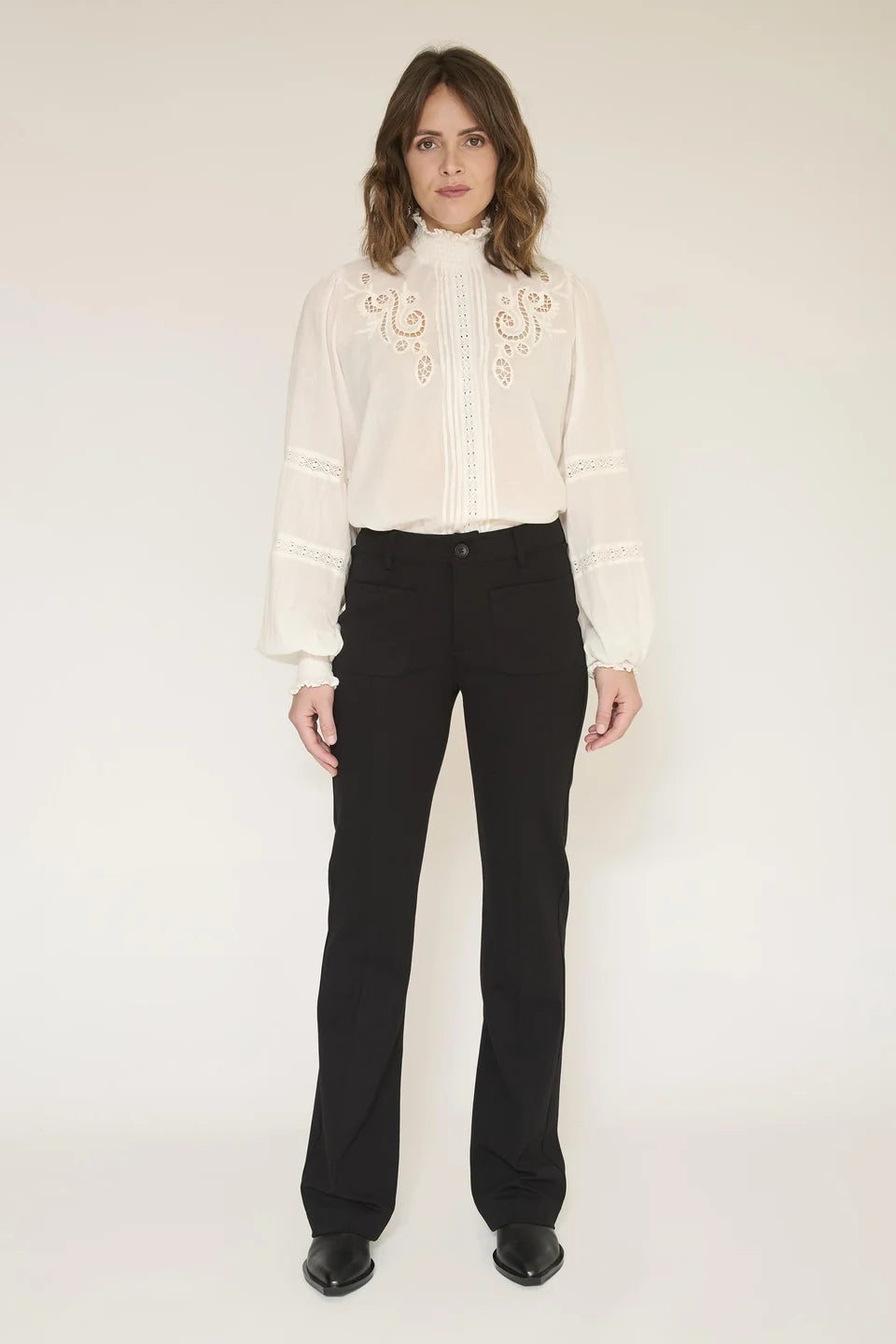 A woman stands against a plain light background wearing a cream long-sleeve blouse with ornate embroidery around the high neck and cuffs, paired with black pants and black shoes.