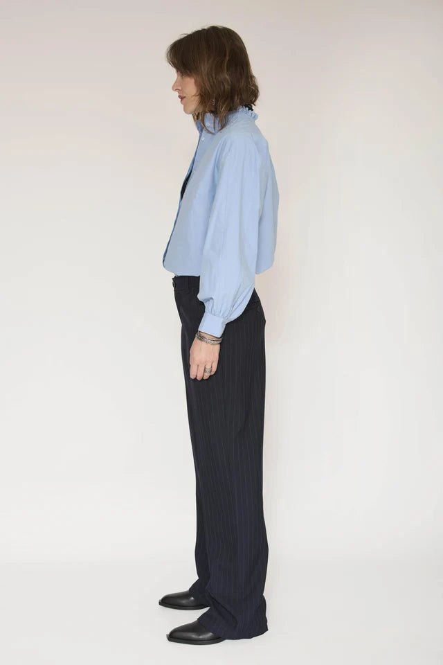 A woman shown in profile wearing a light blue button-up shirt, dark pinstriped trousers, and black shoes against a plain light background.