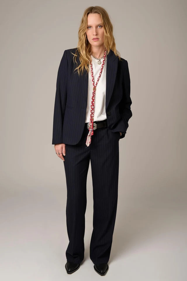 A model stands against a neutral studio background wearing a dark navy pinstripe suit with a white shirt, a long red-and-white patterned scarf, and black shoes.