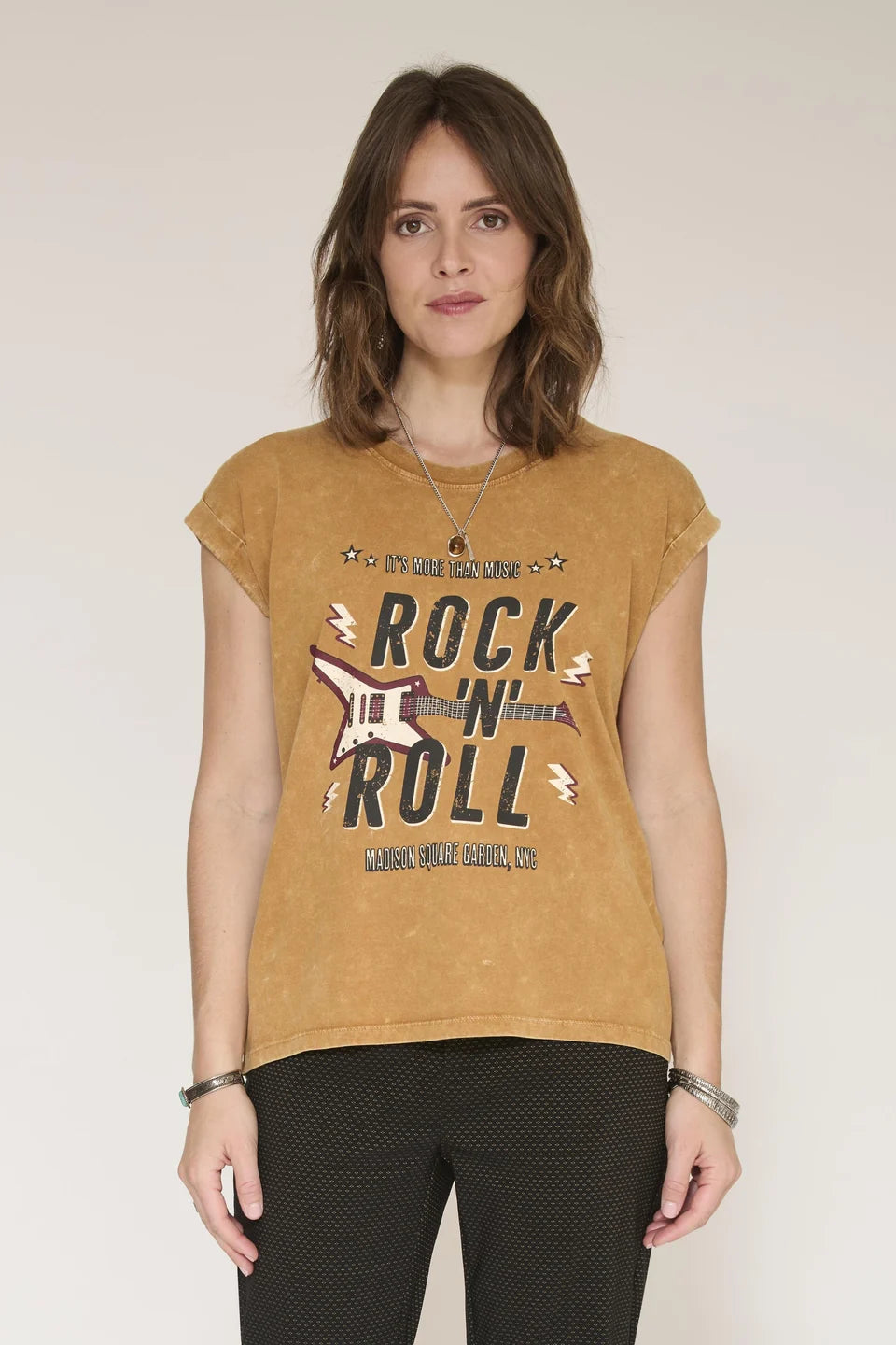 A woman modeling a cropped, faded mustard yellow t-shirt with a distressed graphic that reads 'ROCK N' ROLL' and 'LEONARDO VALLANCE, CO'. She is wearing black patterned pants against a plain, light beige studio background.