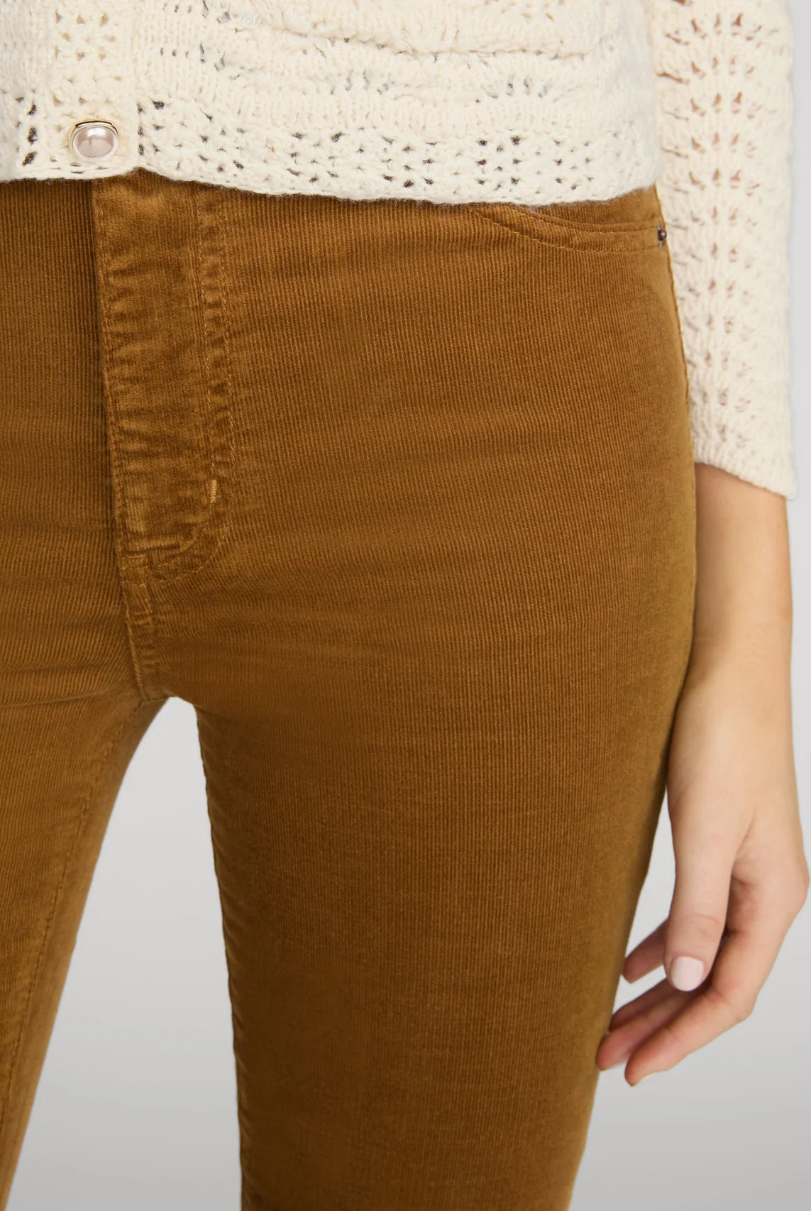 Close-up of a person wearing brown corduroy pants and a cream knit cardigan, cropped from the chest down against a light gray studio background.