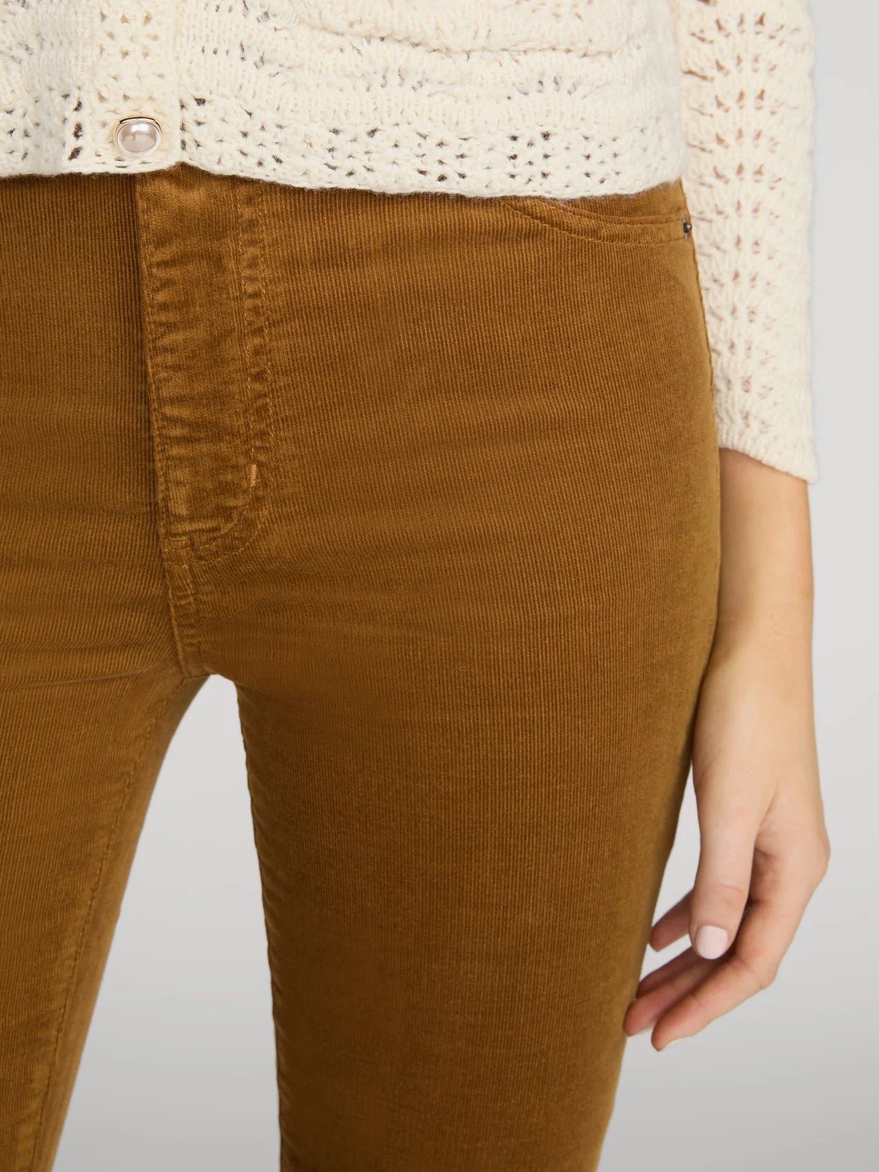 Close-up of a person wearing brown corduroy pants and a cream knit cardigan, cropped from the chest down against a light gray studio background.