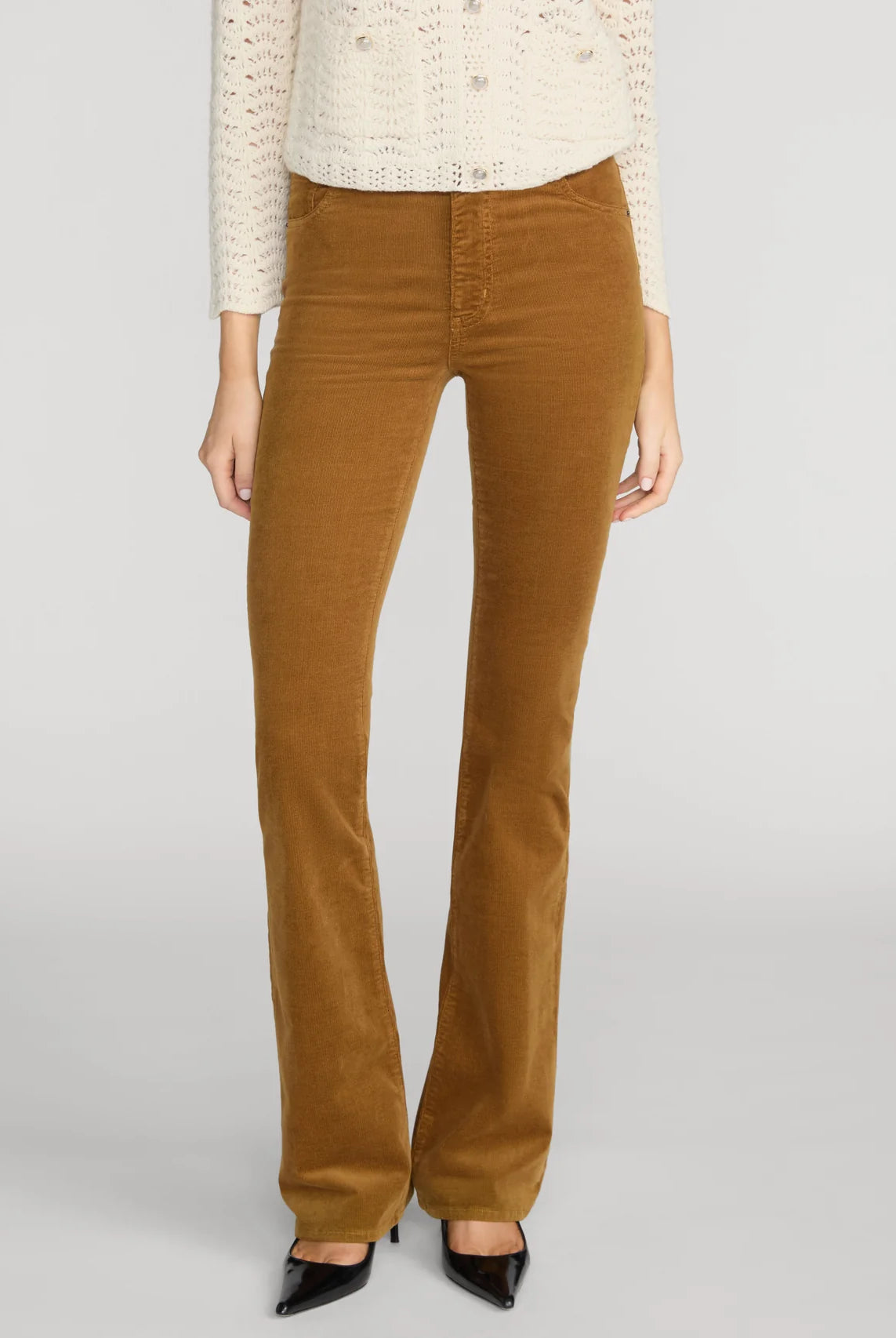 A model wearing a pair of brown corduroy flare pants, styled with a cream knit sweater and black pointed-toe heels, against a light gray studio background.