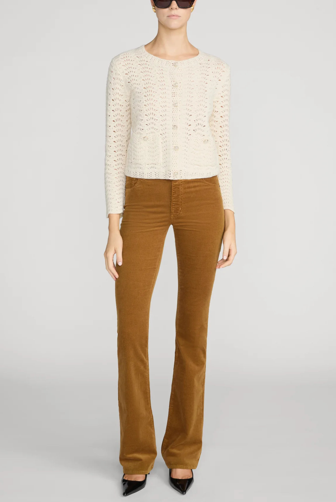 A model wearing a cream-colored textured knit cardigan, brown corduroy flare pants, and black pointed-toe heels, photographed against a neutral light gray studio background.