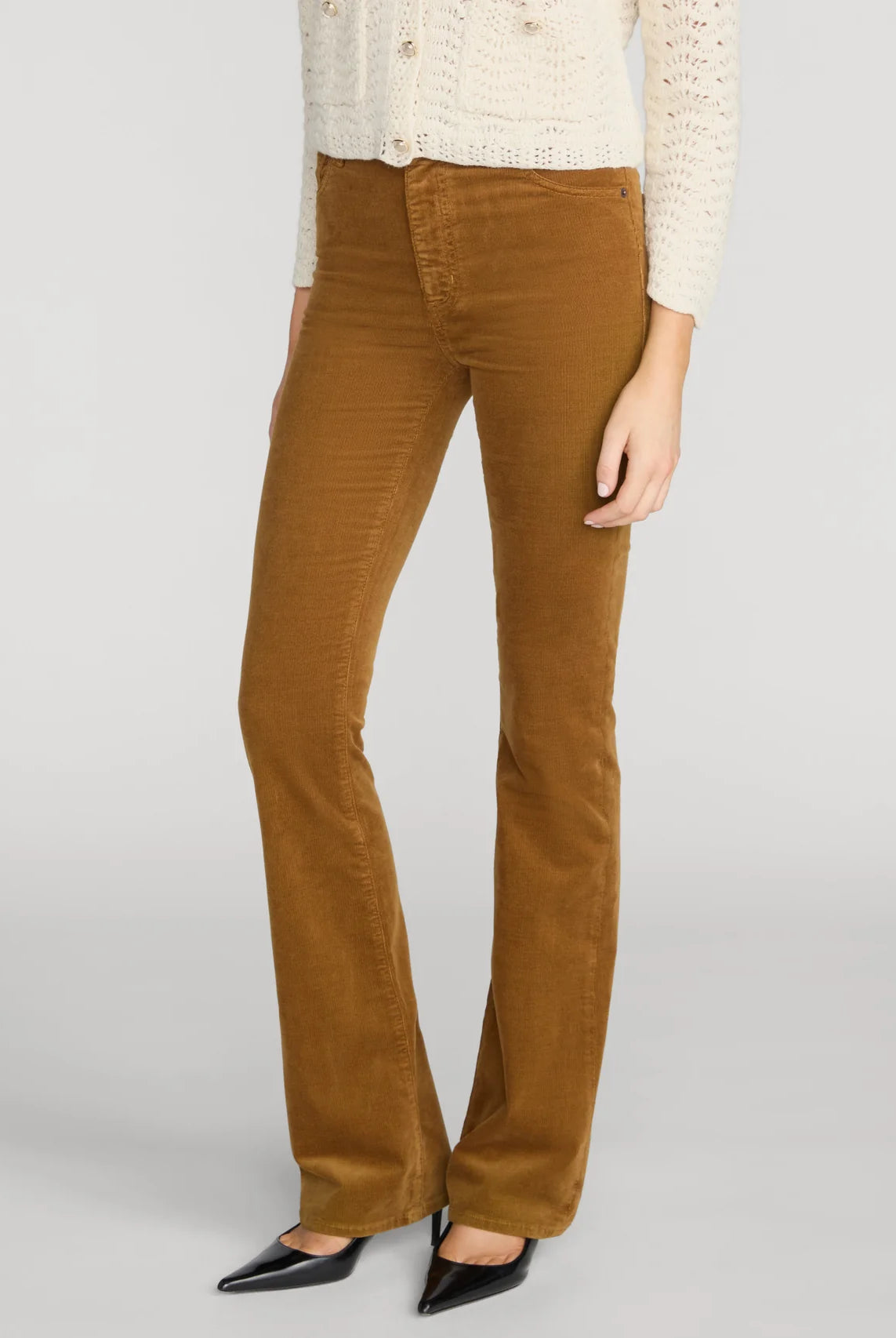 A model wearing a pair of brown corduroy flare pants, styled with a cream knit cardigan and black pointed-toe flats, against a light gray studio background.
