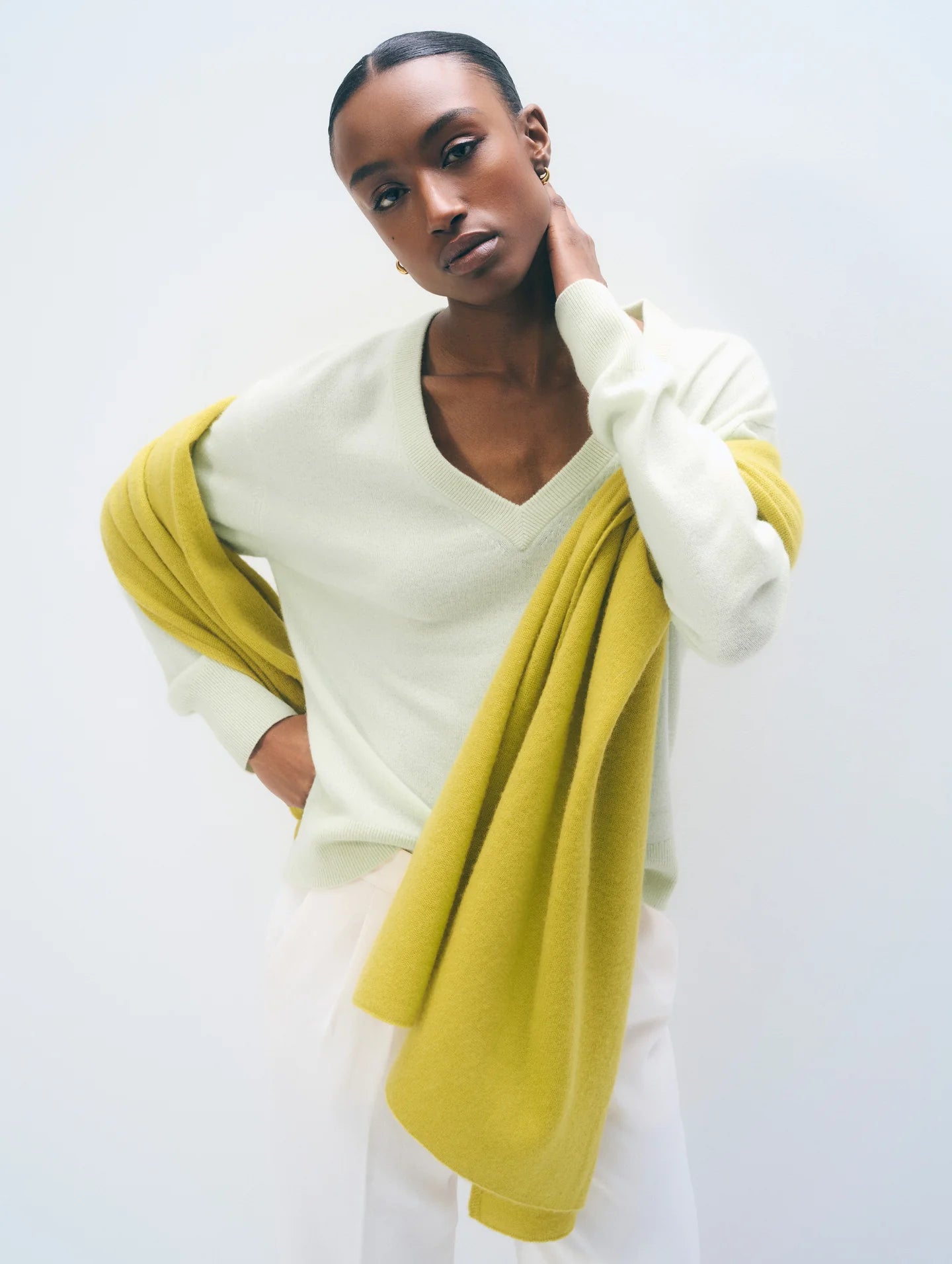 A model wearing a bright yellow-green scarf over a light green V-neck sweater and white trousers, standing against a plain white studio background.