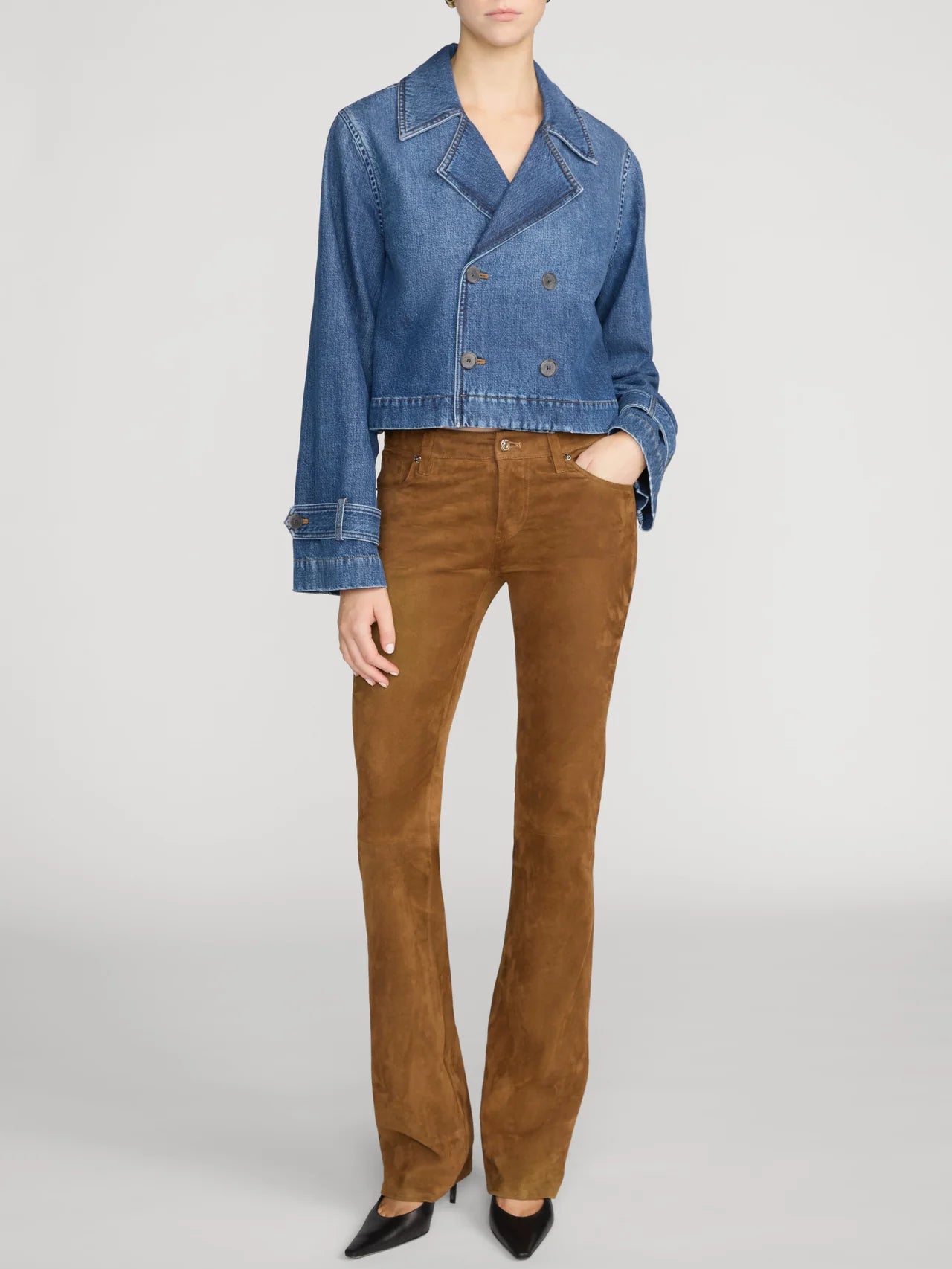 A model wearing a cropped blue denim jacket and brown suede flare pants, paired with black pointed-toe heels, against a light gray studio background.