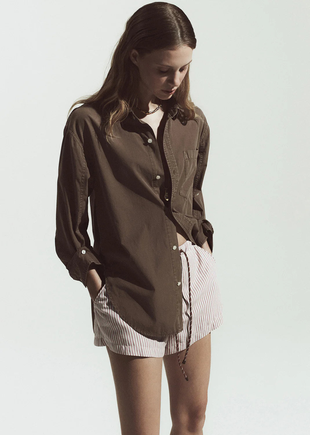 Woman wearing a brown shirt over a white top with pink shorts on a plain background