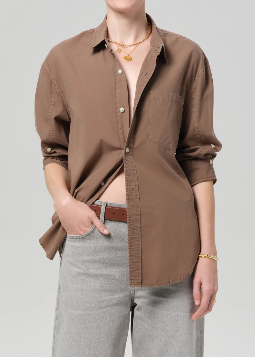 Person wearing a brown shirt over a tank top with light gray pants on a plain background