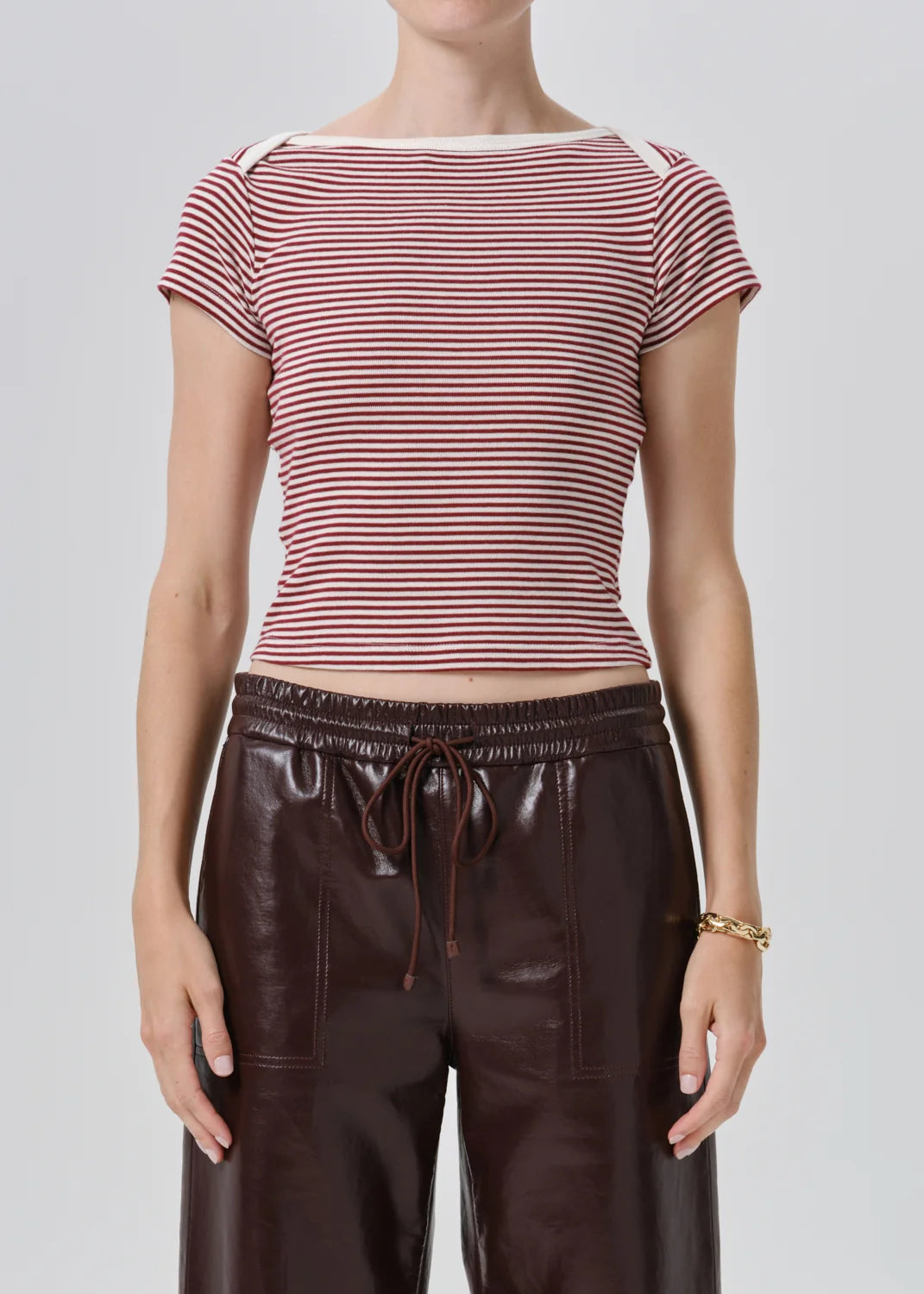 A model wearing a short-sleeve striped knit top with red and white horizontal stripes, paired with dark brown faux-leather pants, against a light gray studio background.