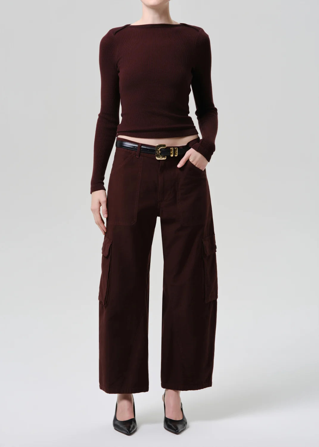 A model wearing a dark brown long-sleeve knit top and matching dark brown wide-leg cargo pants, styled with a black leather belt and black pointed-toe heels against a light gray studio background.