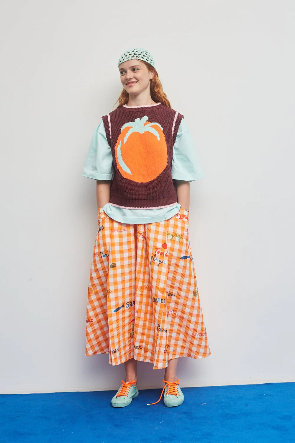 Person wearing a brown sweater with a pumpkin design, light blue shirt, orange checkered skirt, and green sneakers on a white background.
