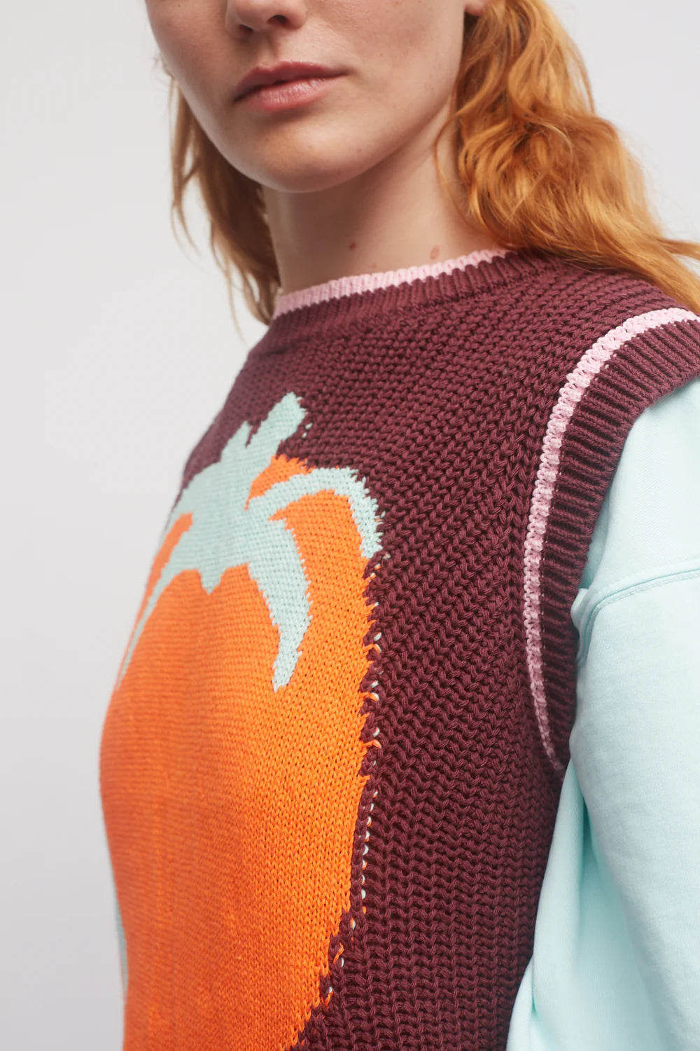 Person wearing a maroon knitted vest with a large orange and green leaf design on a plain background