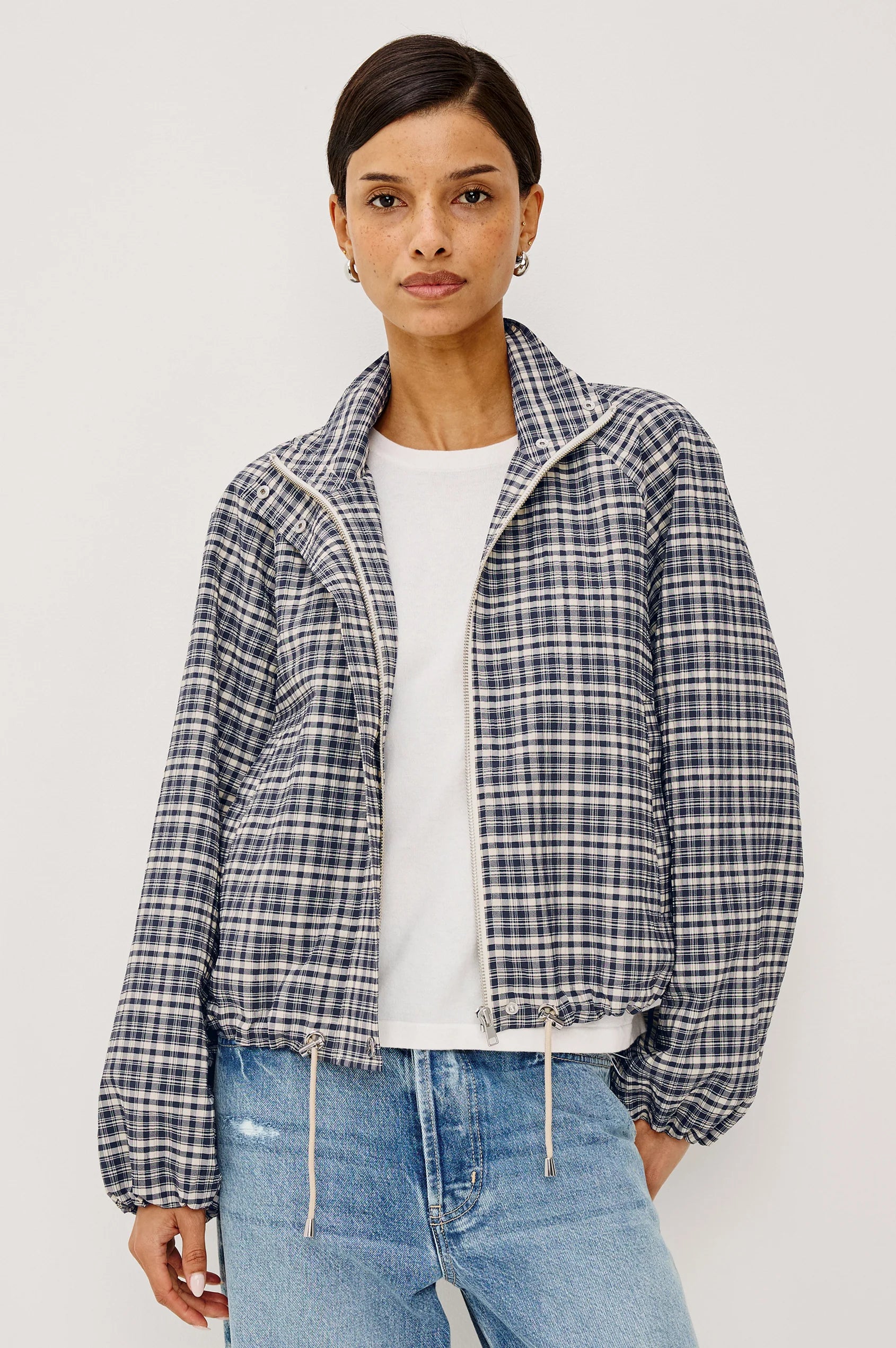 A woman modeling a navy and white checkered long-sleeve cropped jacket with a high stand-up collar, unzipped, paired with light blue jeans, against a plain light gray background.