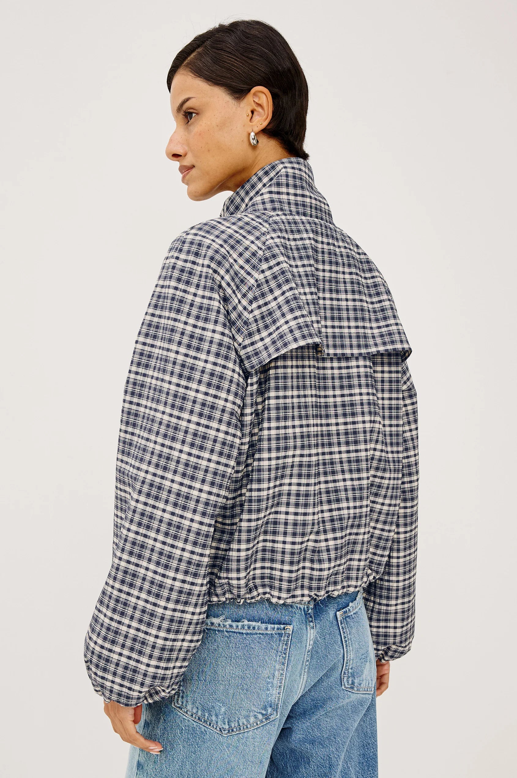 A woman shown from the back wearing a cropped, puffer-style plaid jacket in a gray, white, and black check pattern, paired with blue jeans, against a plain light gray studio background.
