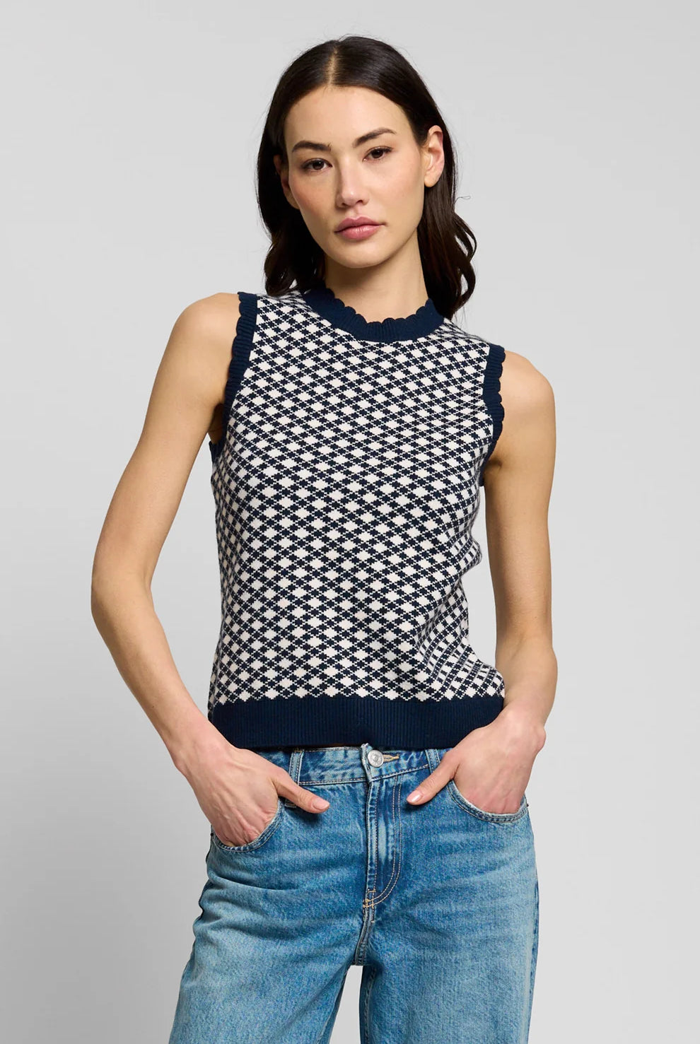 A woman modeling a sleeveless knit top with a navy and white geometric pattern, paired with blue jeans, against a solid light gray background.