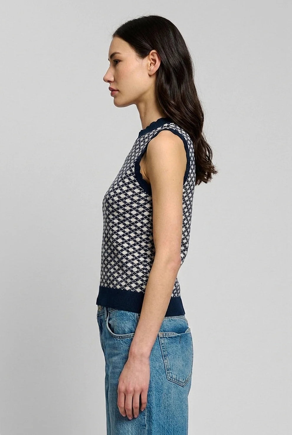 A woman shown in side profile wearing a sleeveless knit top with a navy and white geometric pattern, paired with blue jeans, against a plain light gray studio background.