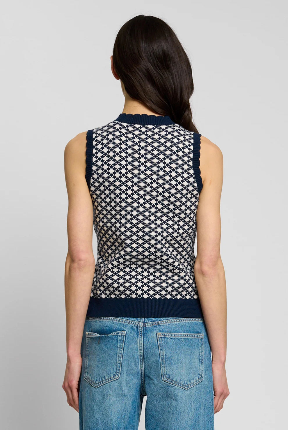 A woman shown in back profile wearing a sleeveless knit top with a navy and white geometric pattern, paired with blue jeans, against a plain light gray studio background.