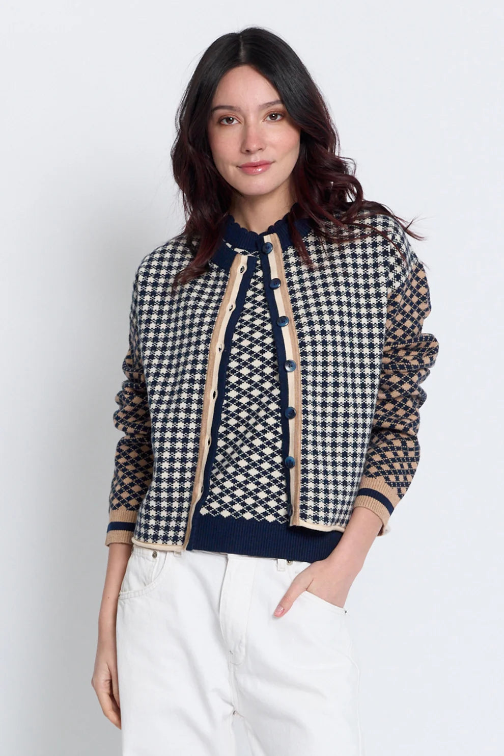 A woman modeling a cropped houndstooth jacket with sheer mesh sleeves, beige trim, and navy buttons, paired with white pants against a light gray studio background.