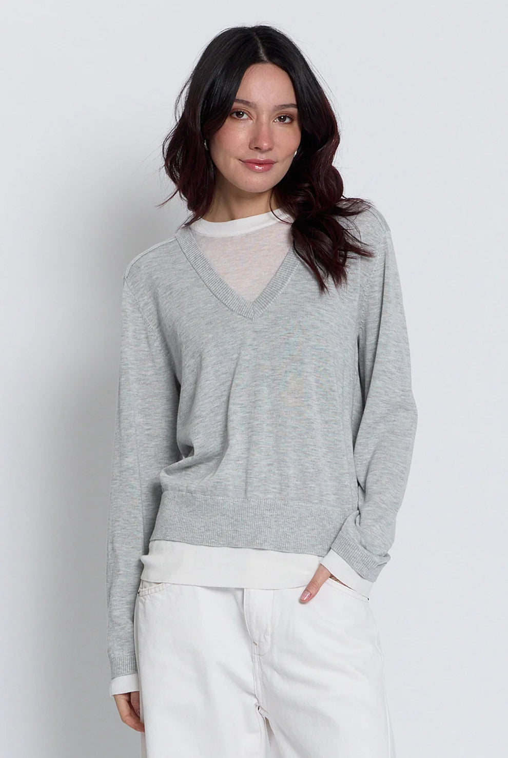 A woman wearing a light gray v-neck knit sweater over a white mesh top, paired with white pants, photographed against a light gray studio background.
