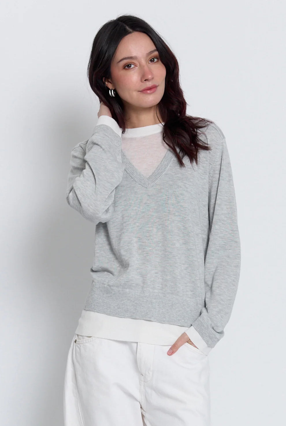 A woman wearing a light gray heathered v-neck sweater over a white mock-neck top and white trousers, standing against a light gray studio background.