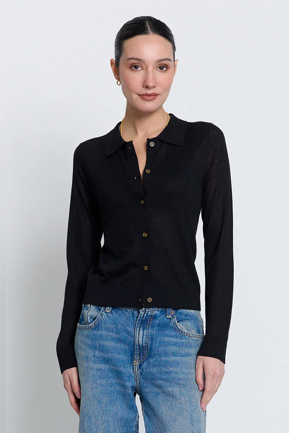 A woman modeling a black long-sleeve knit polo shirt with brown buttons, paired with blue jeans, against a light gray studio background.